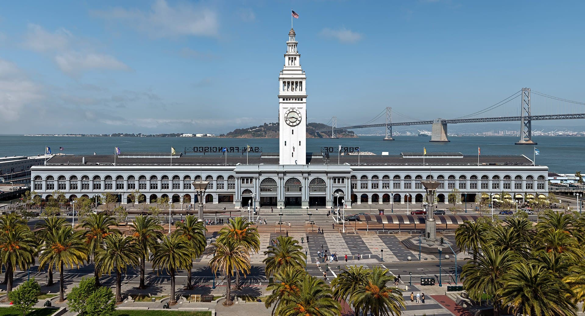 The San Francisco Ferry Building seen from the Embarcadero, its clock tower centred, with the Bay Bridge behind and palm trees along the waterfront.